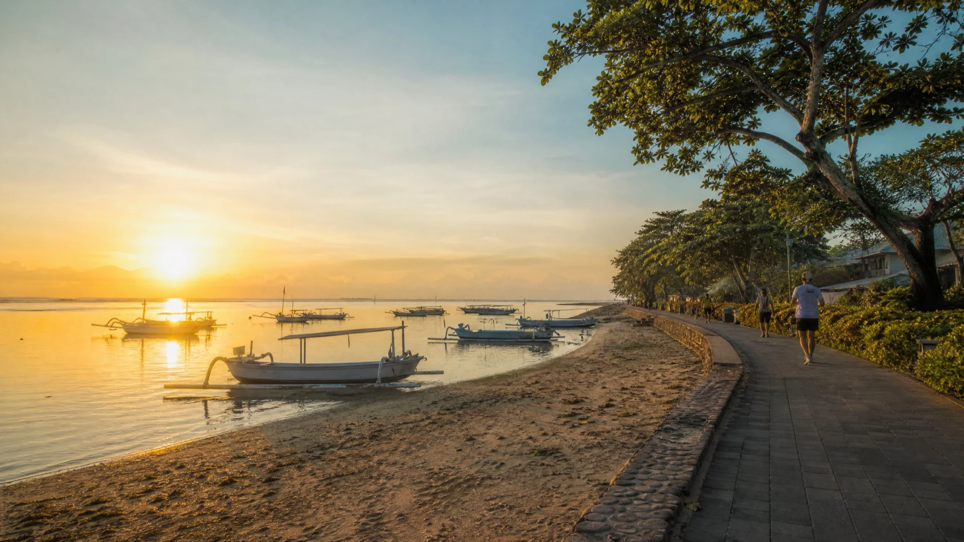 People walking beside traditional boats on Sanur’s calm beachfront promenade at sunrise